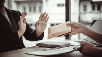 مشهد لمحادثة بين شخصين في مكتب، حيث يرفض أحدهما استلام ظرف بني اللون. يظهر الشخص الذي يقدم الظرف وهو يمد يده، بينما يرفع الشخص الآخر يديه في إشارة إلى الرفض. في الخلفية، توجد نوافذ كبيرة تسمح بدخول الضوء الطبيعي.