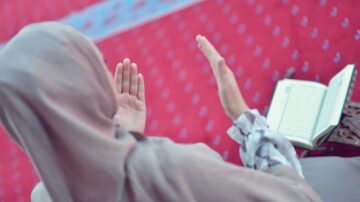 امرأة ترتدي حجابًا، تجلس على سجادة صلاة حمراء وتؤدي الدعاء، بينما كتاب القرآن الكريم موضوع بجانبها.