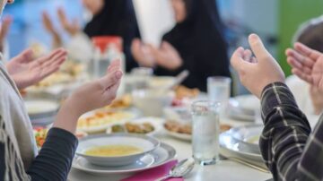 مجموعة من الأشخاص يجلسون حول مائدة طعام، يرفعون أيديهم للدعاء. تحتوي المائدة على أطباق متنوعة من الطعام، منها الشوربة والمقبلات، مع كؤوس من الماء. الأجواء تعكس روح الجماعة والتشارك في وجبة.