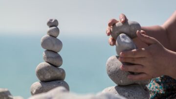 تظهر الصورة شخصًا يكدس الحجارة على شاطئ البحر، حيث تتكون مجموعة من الحجارة المرتبة في شكل عمودي على اليمين، بينما يقوم الشخص بترتيب حجارة أخرى على اليسار. الخلفية تحتوي على مياه زرقاء هادئة.