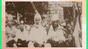 صورة تاريخية لمجموعة من الرجال يجلسون في الخارج، يرتدون ملابس تقليدية. يظهر في الخلفية جدار حجري وأشجار. يبدون جادين وهم يحملون كتبًا، مما يشير إلى اجتماع ثقافي أو ديني.