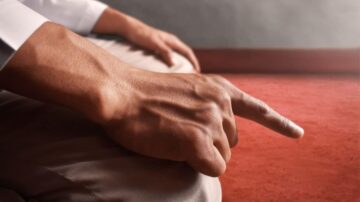 يد تشير بإصبع مستقيم نحو الأرض، مع التركيز على تفاصيل البشرة. تظهر اليد في وضعية الاستراحة على ركبتي شخص يجلس على سجادة حمراء. الإضاءة تضيف عمقًا للصورة، مما يبرز نعومة اليد والملابس.