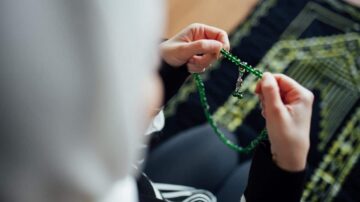 امرأة ترتدي حجابًا، تمسك بسلسلة من الخرز الأخضر، وتجلس على سجادة صلاة. تعكس الصورة لحظة من التأمل والروحانية.