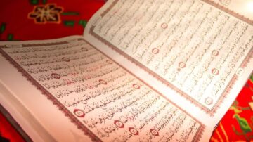 كتاب مفتوح يظهر نصوص عربية من القرآن الكريم مع تزيينات جميلة. يحتل الكتاب مركز الصورة على سجادة ملونة، مما يعكس التراث الثقافي والديني.