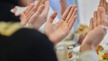 أيدي مرفوعة في دعاء جماعي حول مائدة طعام، مع التركيز على التفاصيل مثل الخواتم. تعكس الصورة الأجواء الروحية والتقاليد الثقافية.