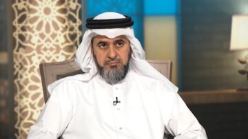 رجل يرتدي الزي التقليدي العربي مع عمامة بيضاء، يجلس في استوديو مع خلفية مزخرفة وإضاءة دافئة، يظهر تعبيره الجاد.