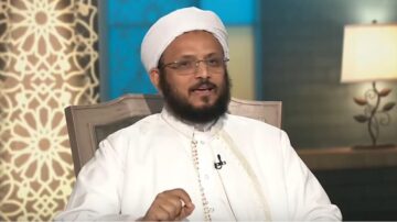رجل يرتدي زيًا تقليديًا إسلاميًا، يجلس في استوديو مع خلفية مزخرفة، يتحدث في برنامج تلفزيوني، يظهر تعبيره الجاد وثقافته الدينية.