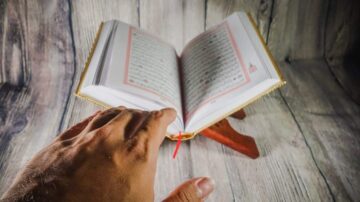 كتاب مفتوح على حامل خشبي، مع يد تشير إلى الصفحات. الخلفية تتكون من خشب بلون داكن.