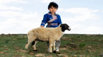 طفل صغير يرتدي زي تقليدي ملون يقف في حقل مفتوح بجانب خروف. السماء زرقاء مع سحب بيضاء، والأرض مغطاة بالعشب والأحجار. تعكس الصورة الحياة الريفية والتراث الثقافي.