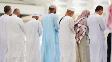 مجموعة من الرجال يرتدون ملابس تقليدية مختلفة، يقفون في صف للصلاة. يظهر في الصورة تنوع الأزياء، بما في ذلك الثوب والشماغ، في بيئة دينية.