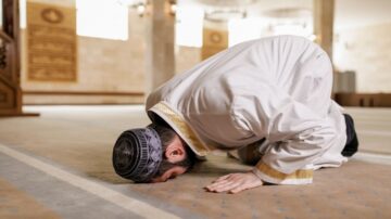 رجل مسلم يؤدي السجود في مسجد، يرتدي زي تقليدي ويظهر التركيز والعبادة في أجواء هادئة.