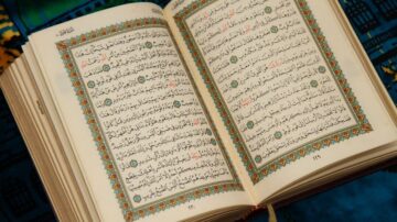 كتاب مفتوح يحتوي على نصوص عربية مزخرفة، يظهر فيه آيات من القرآن الكريم مع تزيينات ملونة حول الحواف. الخلفية تظهر سجادة بتصميم تقليدي.