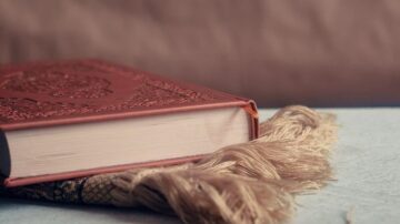 كتاب مزخرف بلون بني، موضوع على سطح خشبي، مع حواف دافئة. يظهر جزء من سجادة مزخرفة باللون البيج بجانب الكتاب، مما يضيف لمسة تقليدية.