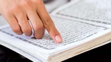 يد تشير إلى نص مكتوب في كتاب، مع صفحات مفتوحة تعرض خطوطاً عربية.