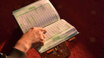 كتاب مفتوح على حامل، مع يد تشير إلى نص مكتوب باللغة العربية. الخلفية حمراء، مما يبرز تفاصيل الصفحات.