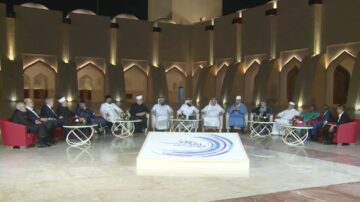 لقاء حواري في استوديو أنيق، يضم مجموعة من الرجال والنساء الجالسين حول طاولة. الخلفية معمارية تقليدية، مع إضاءة دافئة وأقواس. الحضور يرتدون ملابس متنوعة، مما يعكس تنوع الثقافات. الموضوعات المحتملة تشمل القضايا الاجتماعية والدينية.