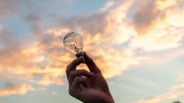 مصباح كهربائي يحمل بيد أمام سماء مزينة بالسحب الملونة في وقت الغسق.