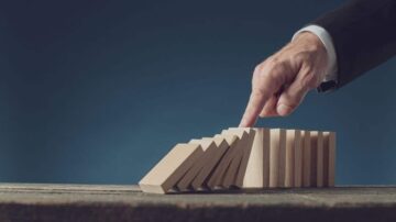يد تظهر إصبعًا يلمس قطعة خشبية من مجموعة من قطع الدومينو المرتبة بشكل مائل، مما يوحي بتأثير الدومينو. الخلفية زرقاء داكنة، وتضفي لمسة احترافية على المشهد.
