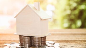 منزل خشبي صغير موضوع على كومة من العملات المعدنية، مما يرمز إلى مفهوم الاستثمار العقاري والمال. خلفية ضبابية تبرز جمال الصورة.