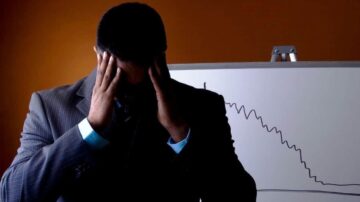 رجل يرتدي بدلة رسمية، يُظهر تعبيرات القلق والإحباط، يضع يديه على رأسه. خلفه، لوحة بيضاء تُظهر رسمًا بيانيًا منحنيًا للأسفل، مما يشير إلى تراجع أو خسائر.