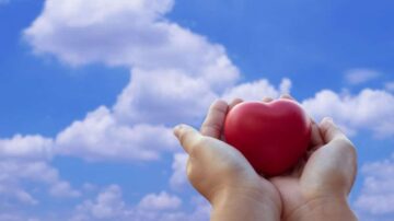 يد تحمل قلبًا أحمر تحت سماء زرقاء مزينة بسحب بيضاء. تعبر الصورة عن الحب والأمل.