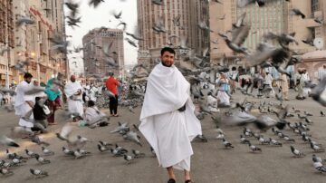 رجل يرتدي الإحرام يقف وسط حشود من الحجاج في مكة المكرمة، تحلق حوله أسراب من الحمام. تظهر المباني العالية في الخلفية، مما يبرز أجواء الحج.