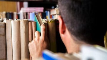 رجل يختار كتابًا من رفوف مكتبة مليئة بالكتب، يظهر جزء من يده وقبضة الكتاب، مع التركيز على بيئة القراءة والمطالعة.
