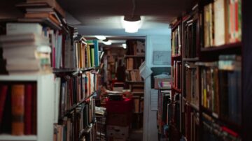 مكتبة قديمة تحتوي على صفوف من الكتب المكدسة على الرفوف. الإضاءة خافتة، مما يخلق جوًا هادئًا. تظهر تفاصيل الكتب بألوان وأحجام متنوعة، مع ممر ضيق بين الرفوف.