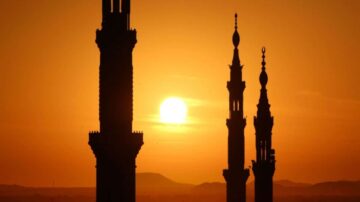 غروب الشمس خلف مآذن مساجد، مع ظلال داكنة تظهر تفاصيل المآذن في السماء البرتقالية.