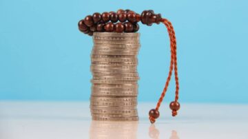 عملات معدنية مرتبة في عمود مع سبحة خشبية باللون البني، خلفية زرقاء. العملات تعكس مفهوم الادخار والاستثمار، بينما تعبر السبحة عن الروحانية والتركيز.