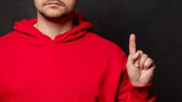 رجل يرتدي سترة حمراء، يرفع إصبعه السبابة في إشارة إلى التوقف أو الانتباه، مع خلفية داكنة.