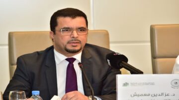 رجل يرتدي بدلة رسمية يتحدث في مؤتمر، يجلس أمام ميكروفون وزجاجة ماء. خلفه لافتة تحتوي على نص عربي، مما يشير إلى حدث رسمي أو حلقة نقاش.