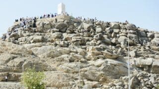 Group atop rocky hill of arafa with white monument under clear sky, sparse vegetation visible.