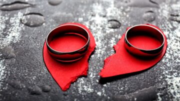 خواتم زفاف فضية موضوعة على قلبين أحمرين ممزقين، مع قطرات ماء على سطح داكن، تعبر عن موضوع الحب والفراق.