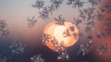 ثلوج متجمدة تتلألأ أمام خلفية ضبابية لشمس غاربة، مع ألوان دافئة تتداخل في السماء.