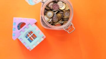 صورة لوعاء زجاجي يحتوي على عملات معدنية متنوعة، بجوار نموذج بيت ملون بلونين. الخلفية برتقالية.