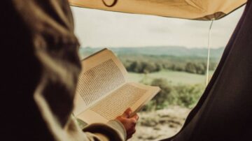 شخص يجلس داخل خيمة ويقرأ كتابًا، مع منظر طبيعي خلاب من خلال فتحة الخيمة، يظهر الحقول والتلال في الخلفية.