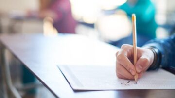 يد تظهر وهي تمسك بقلم رصاص أثناء كتابة على ورقة اختبار، مع خلفية غير واضحة لطلاب آخرين في فصل دراسي.