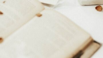 كتاب مفتوح يظهر صفحات غير واضحة مع تفاصيل غامضة، محاط بأوراق ومكونات أخرى، يخلق جوًا من القراءة والتأمل.