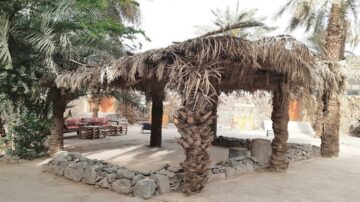 كوخ مصنوع من سعف النخيل محاط بأشجار النخيل وحجارة في الأرض. يظهر في الخلفية أثاث خارجي مخصص للجلوس، مما يخلق جوًا هادئًا في منطقة طبيعية.