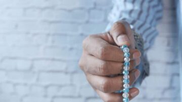 يد تحمل سوارًا مكونًا من كرات زجاجية ملونة باللونين الأزرق والشفاف، مع خلفية من جدار من الطوب الأبيض.