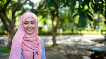 امرأة ترتدي حجابًا ورديًا، مبتسمة في حديقة ذات أشجار خضراء، مع خلفية مشوشة تبرز جمال الطبيعة.