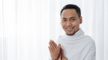 رجل مبتسم يرتدي لباساً أبيض ويعقد يديه معاً في وضع الصلاة، يقف أمام ستائر بيضاء تعكس أجواء هادئة وسلمية.