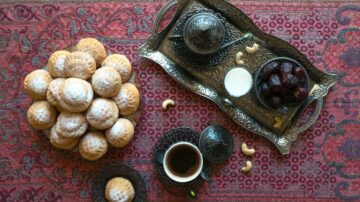 صورة لطبق من الكعك المرشوش بالسكر بجانب صينية فضية تحتوي على الشاي وفواكه مجففة وكاجو. الخلفية سجادة مزخرفة بألوان دافئة.