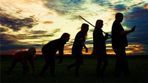 People in a field at sunset in an arrangement similar to the stages of human evolution, the latter carrying a telephone.