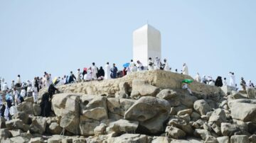 جبل الرحمة في عرفة خلال موسم الحج، حيث يتجمع الحجاج في ملابسهم البيضاء للصلاة والدعاء، مع وجود صخور كبيرة في المقدمة ونصب تذكاري في الأعلى. السماء صافية والجو يعكس روح العبادة والإيمان.