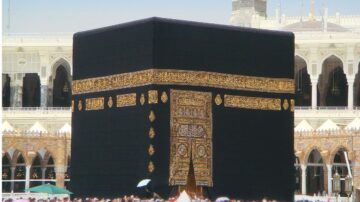 الكعبة المشرفة في مكة المكرمة، مبنى مكعب مغطى بكسوة سوداء مزينة بكتابات ذهبية، تحيط بها مجموعة من الحجاج. تعتبر الكعبة نقطة مركزية في الإسلام ووجهة للحج.