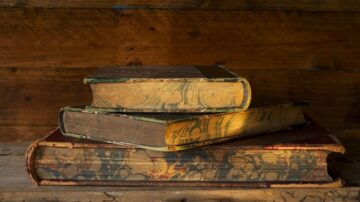 كتب قديمة موضوعة فوق بعضها البعض، مع تغليف مزخرف وألوان دافئة، على سطح خشبي. تعكس الإضاءة تفاصيل الغلاف وملمس الورق.