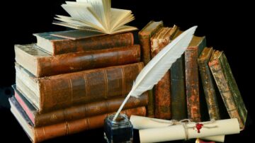 كتب قديمة مكدسة بشكل فني مع كتاب مفتوح في الأعلى، وريشة للكتابة داخل حبر، ولفافة ورقية مربوطة بخيط، تعكس جمال الأدب والتراث الثقافي.