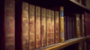 كتب قديمة مرتبة على رف خشبي، تظهر أغلفة مزخرفة بألوان دافئة. تعكس الإضاءة الخافتة جوًا تاريخيًا ورومانسيًا، مما يبرز تفاصيل الكتب.